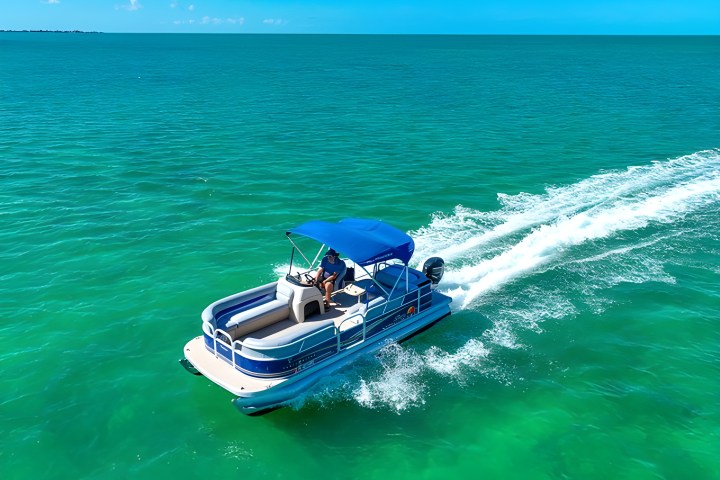 Person driving a blue pontoon boat on clear turquoise water under a bright blue sky.