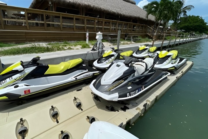 Several jet skis are docked at a marina with a thatched-roof building in the background.