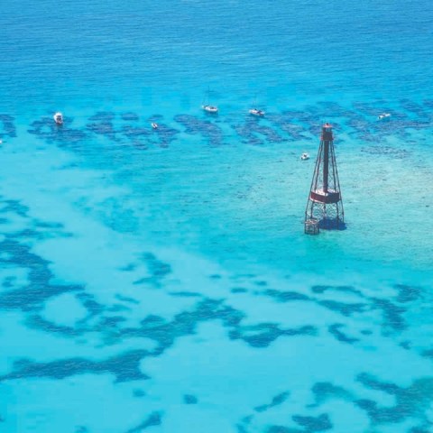 Aerial view of a lighthouse in turquoise ocean with coral patterns and small boats nearby.