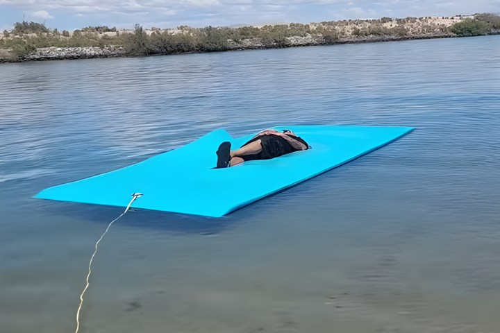 Person relaxing on a floating blue mat in calm water under a clear sky.